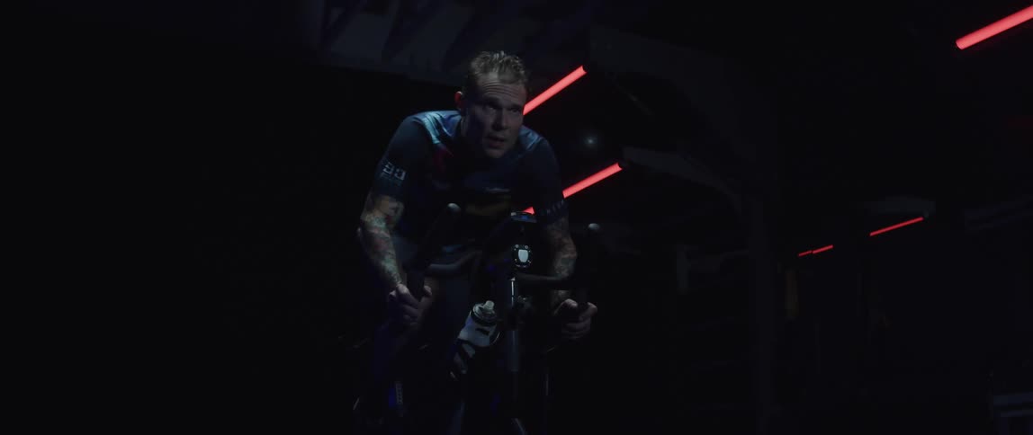 Determined male cyclist with jersey shirt and tattoos rides exercise bike