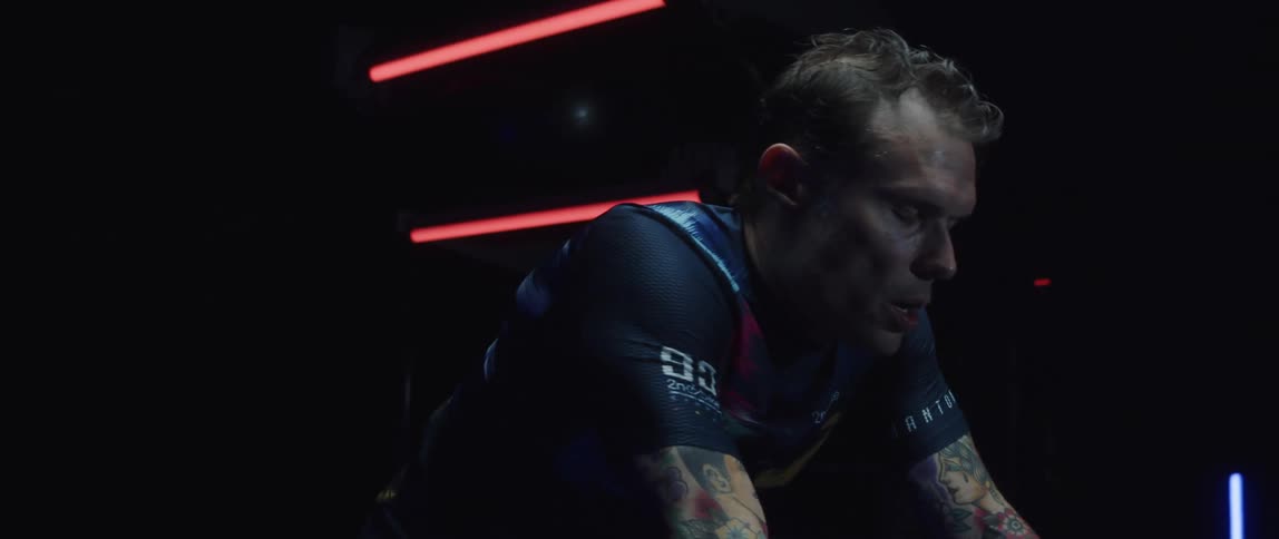 Focused sweaty athletic man exercising on a stationary bike next to a mirror in a dark gym room
