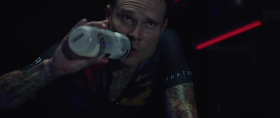 Tattooed man riding a static bicycle indoors drinks water from a plastic bottle