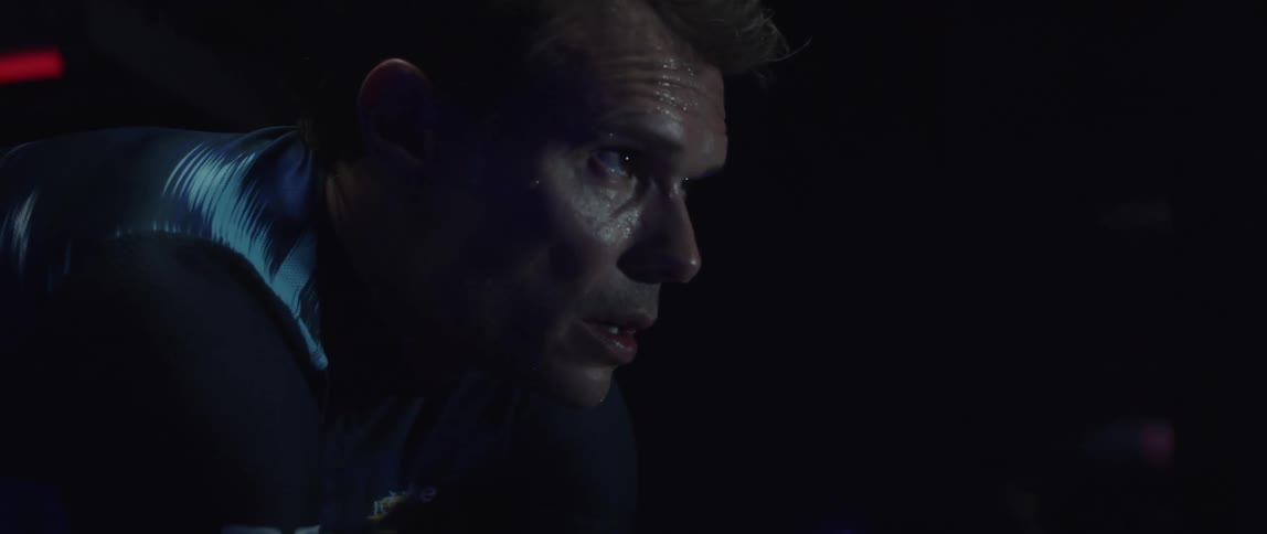 Focused sweaty cyclist man wearing cycling jersey exercising on a stationary bike on a dark room