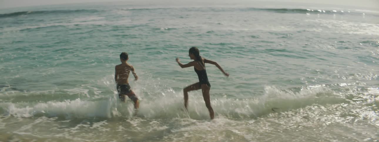 A young girl and boy kick water at each other at the beach before the boy pulls the girl into the water
