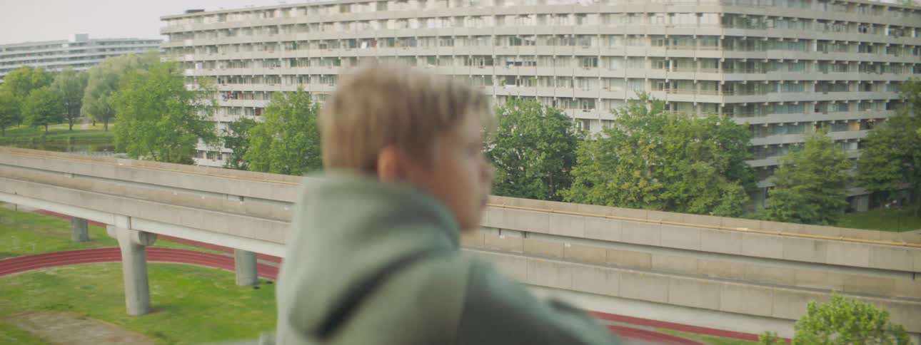A boy with a hoodie standing on a balcony with a running track buildings the sky and clouds in the background