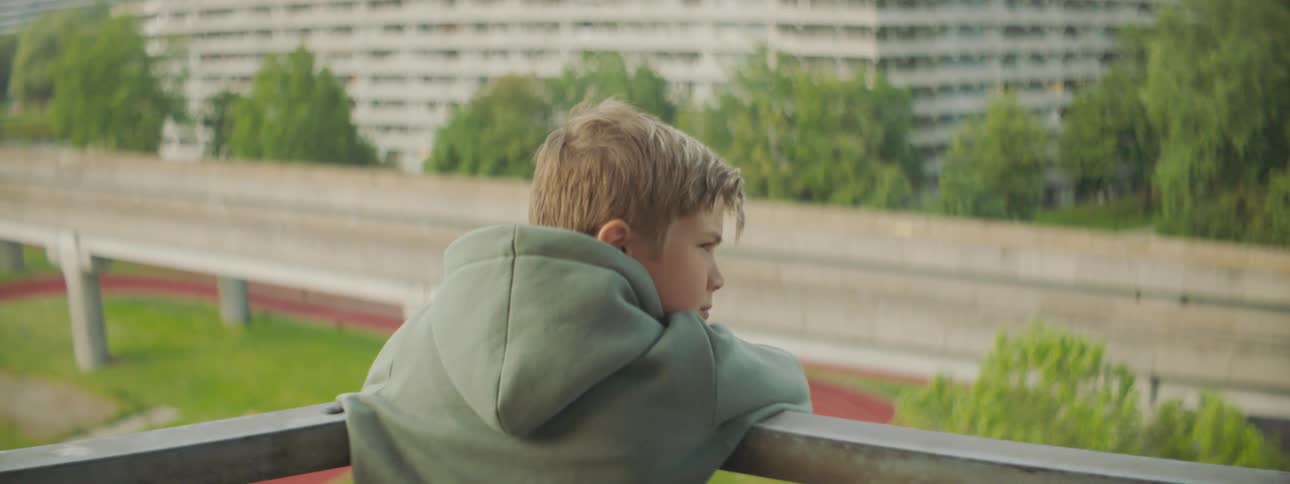 A boy leans against a balcony overlooking a freeway with trees and a building in the background
