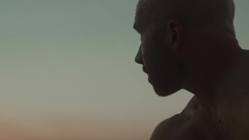 A man outside without a shirt on looks up at the sky as the sun sets