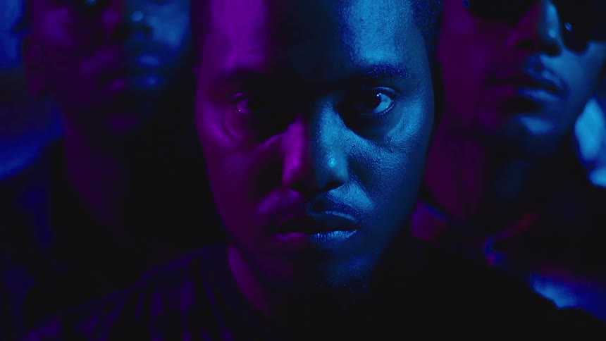 A black man is standing in the darkness with purple and blue light reflecting on his face and there are other men behind him