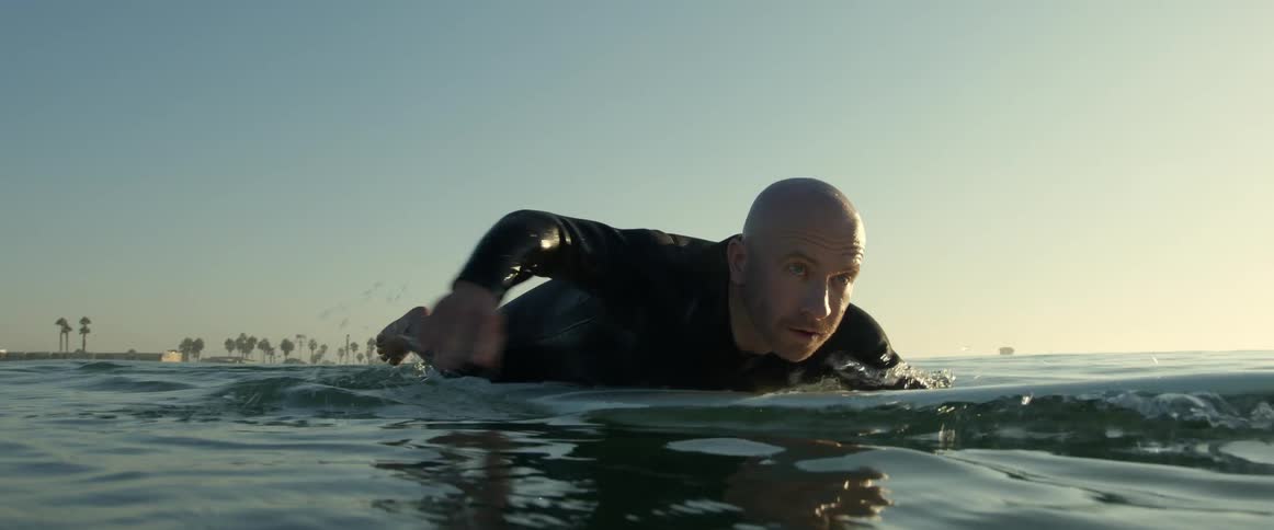 A surfer wearing a black wetsuit lies on his surfboard and paddles with his hands
