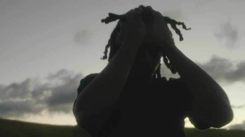 A stressed man runs his hands through his hair while standing outdoors on a hillside in evening