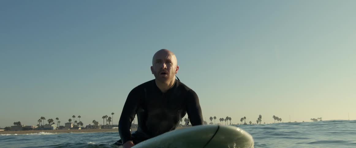 An older white man is sitting on his surf board on the ocean