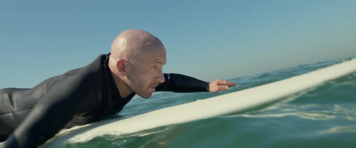An older white man is surfing on the beach in the middle of the day