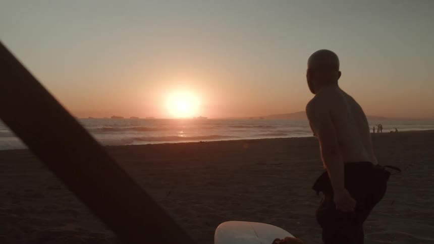 A shirtless man lays a surfboard on the ground and looks out across the ocean at the setting sun