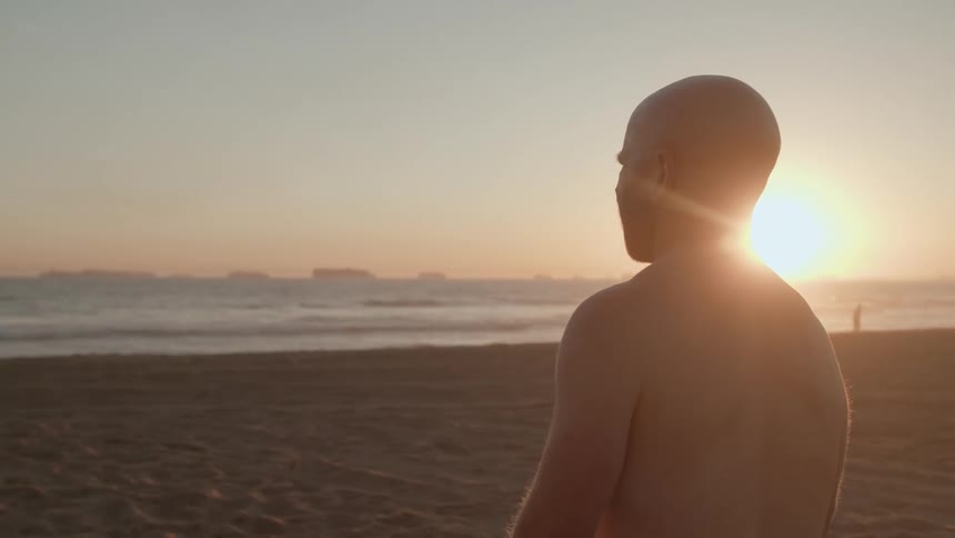 A shirtless man looks out over the ocean while standing on a beach at sunset