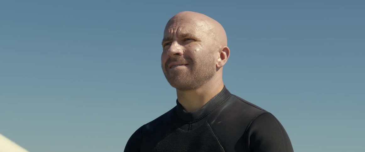 An older white man is sitting on a surf board outside in the middle of the day