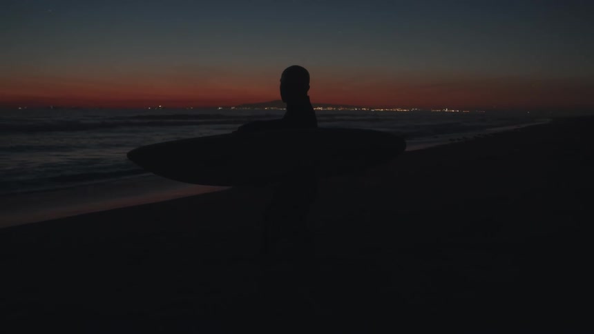 A silhouetted surfer carrying a surfboard on a dark beach at dusk