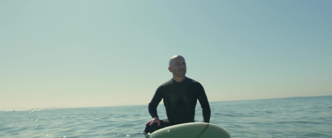 A bald man in a wet suit floating on a surfboard in the ocean