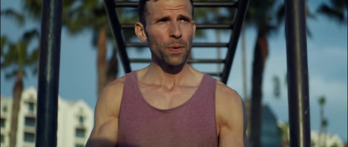 A man in a tank top breathing heavily beneath monkey bars outside
