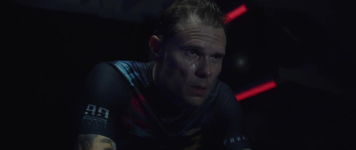 Focused and sweaty male cyclist wearing a cycling jersey exercising on a stationary bike in a dark gym room