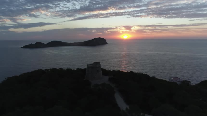 The yellow sun glowing on the horizon above the ocean with a stone tower on a cliff overlooking an island