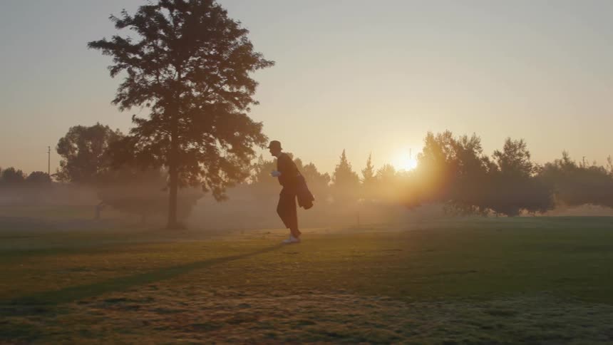 A man wearing a hat and carrying a coat is walking across a field with trees and the sun is shining brightly in a hazy sky