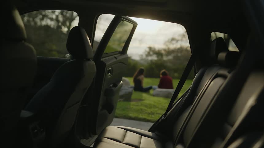 A man and woman sit in a grassy field outside a car and talk