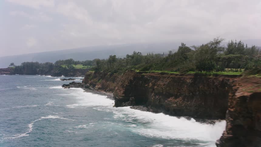 Waves roll into shore below the cliffs along the coast under a cloudy sky