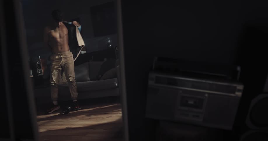 A young man is sitting down lifting a dumbbell and then he puts on a jacket and practices break dancing moves