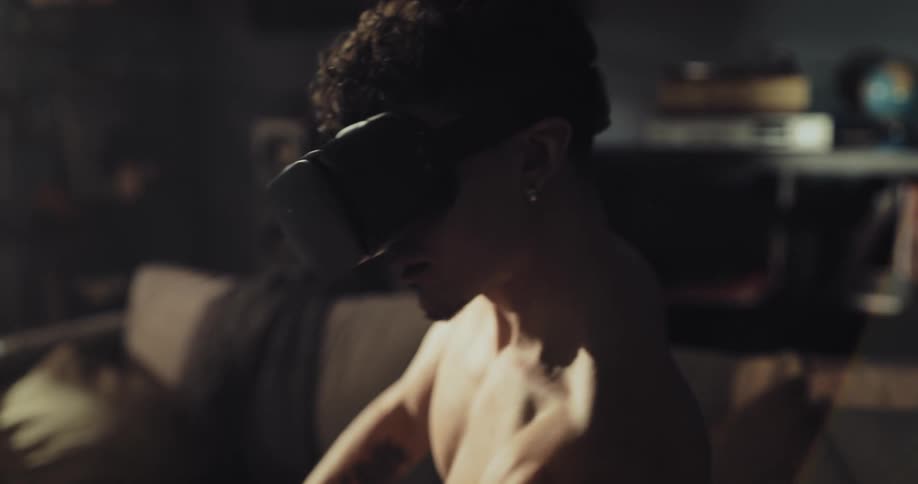 A man wearing a virtual reality headset is also using a dumbbell to work out as he sits on a couch