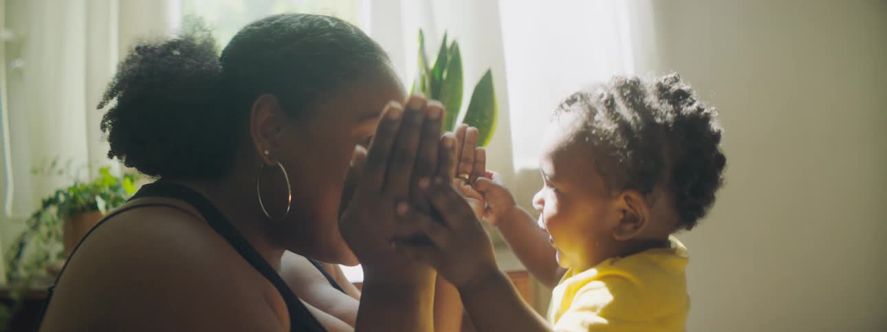 A woman holds her hands over her face as a child tries to remove her hands as they play peek a boo in a room