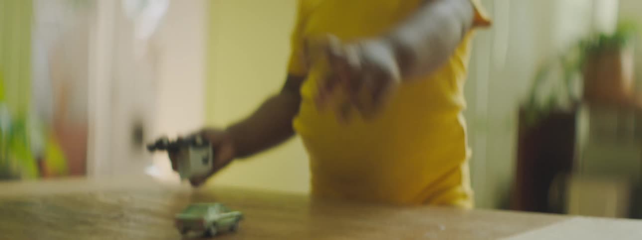 A baby stumbles over to a table where a toy car is spinning and they pick up the car