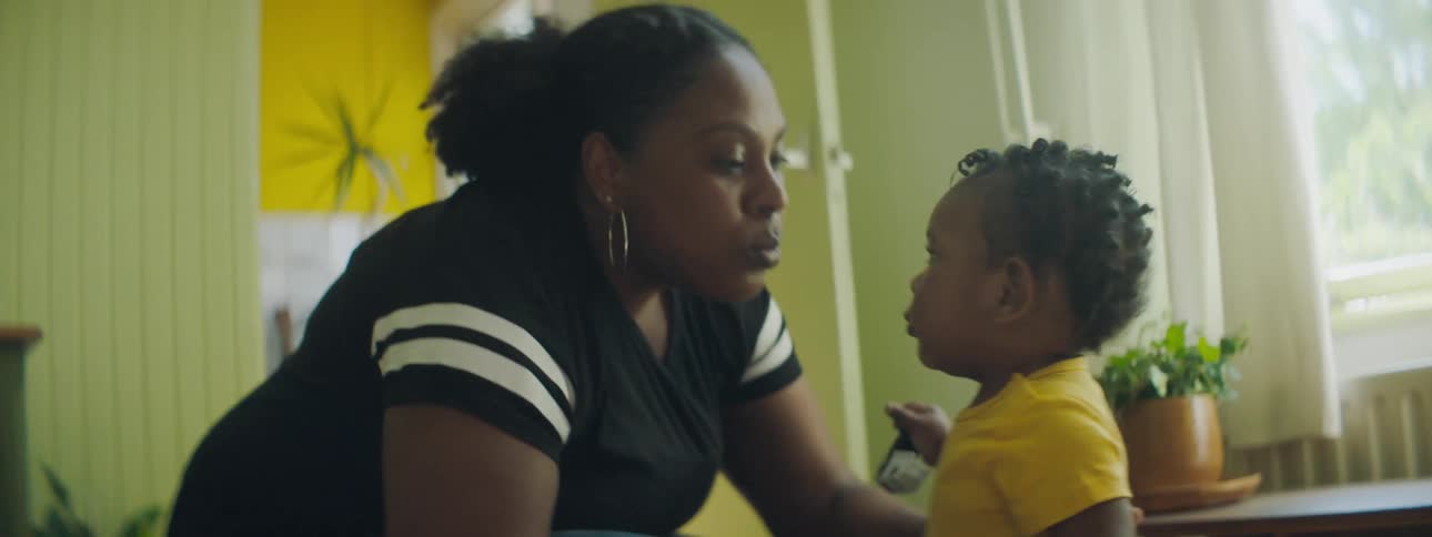 A mother kisses her baby girl who then walks across the room