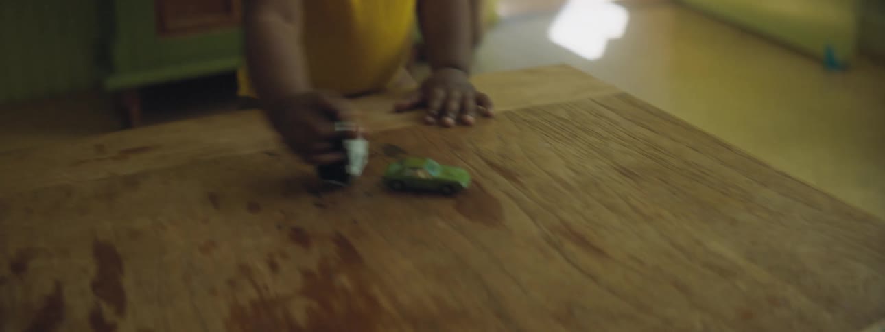 A mother watches as her baby walks to a table and plays with a car