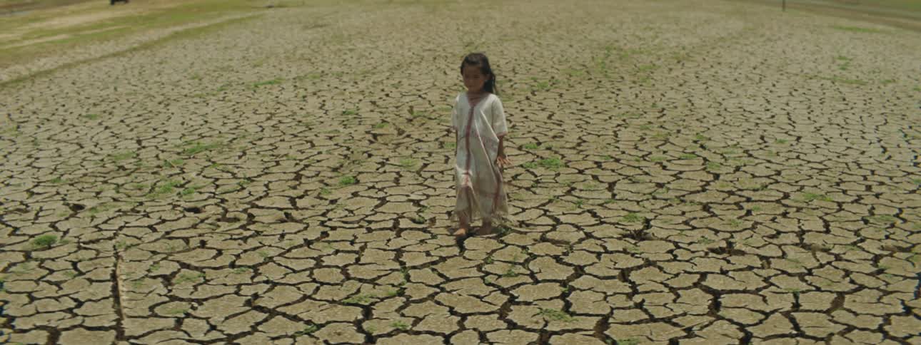 A young girl with black hair and a white dress is walking on dry cracked scorched land