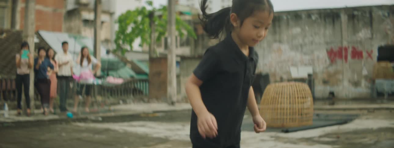 Boy and girl play hopscotch in square near buildings as people watch