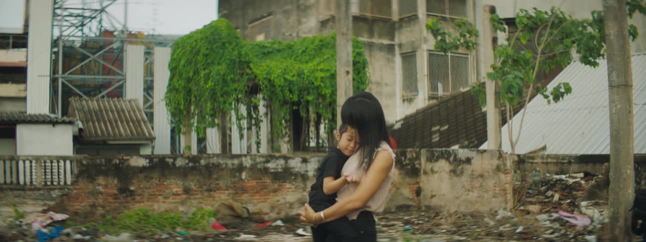 A mother holds her daughter who plays with her hair as they stand outside shabby buildings in a village