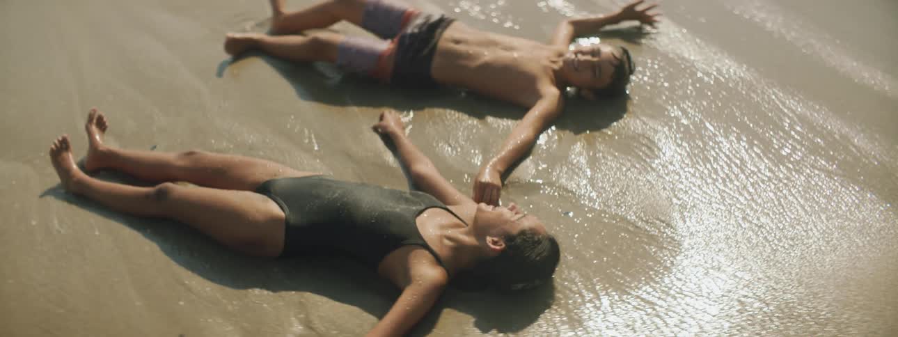 A boy and girl lie on the beach and talk and laugh as waves roll up to them