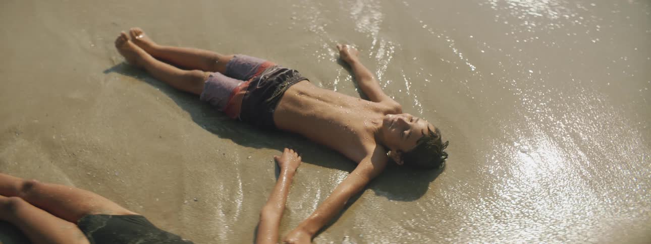A brother and sister lie on the sand and laugh as the water rolls over their bodies