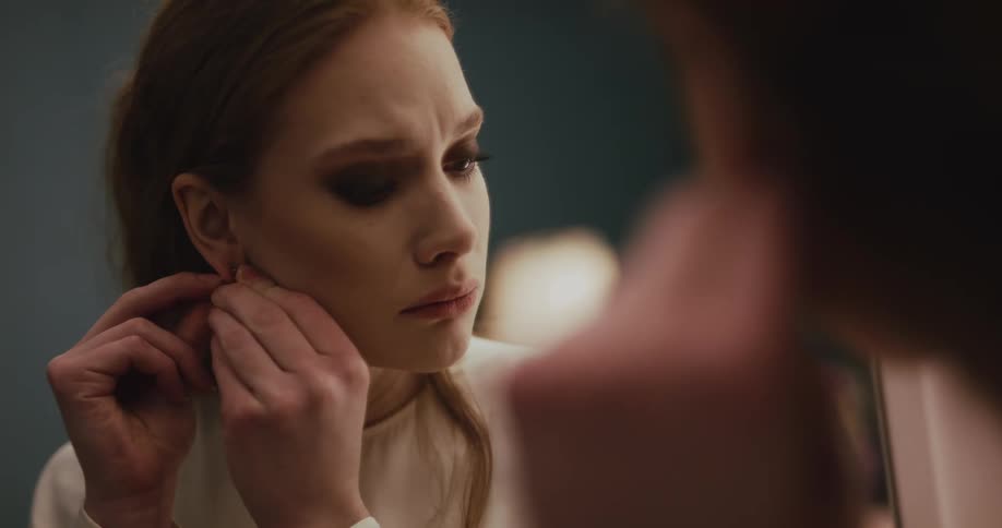 A woman is looking in the mirror fixing her earring and her hair and she is very sad trying not to cry