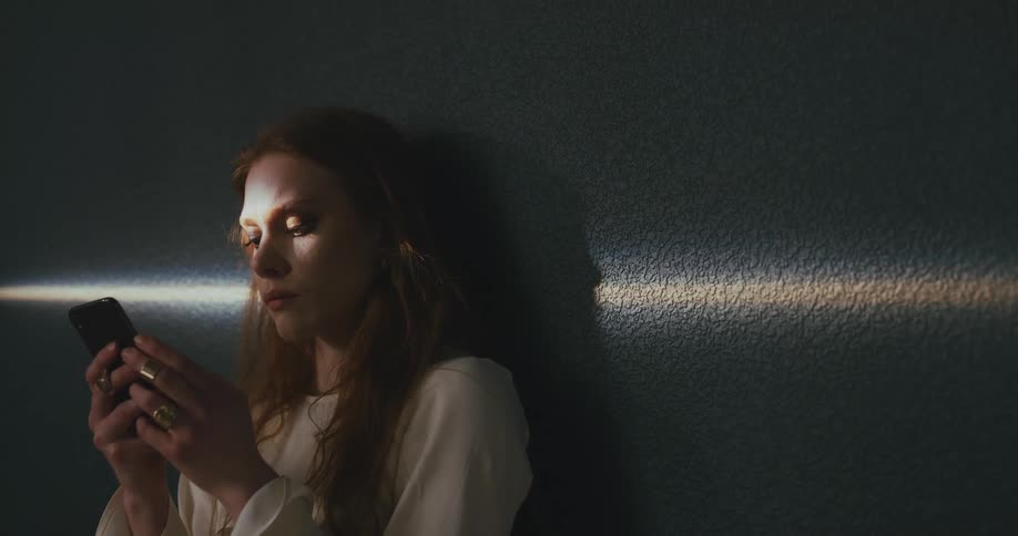 A woman appears bored while looking through her smartphone while leaning against a wall