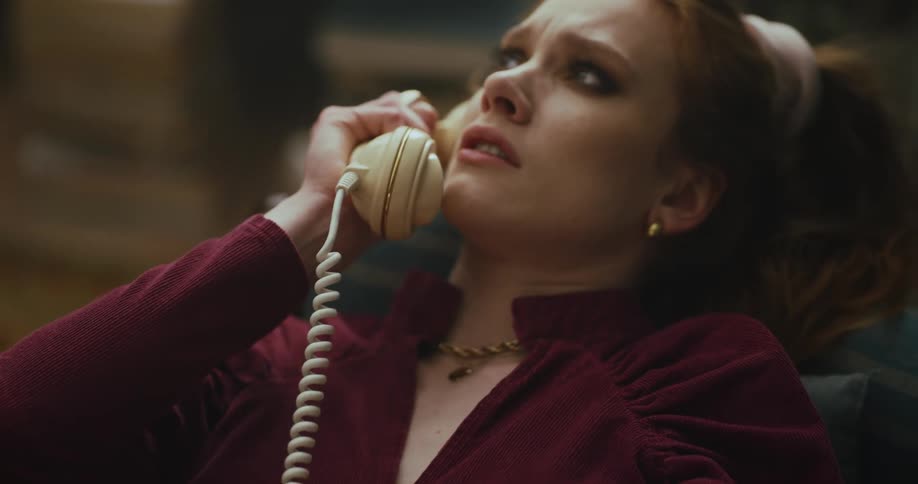 A young woman sits at home and talks on a rotary phone and slowly drops the phone with an expression of sadness
