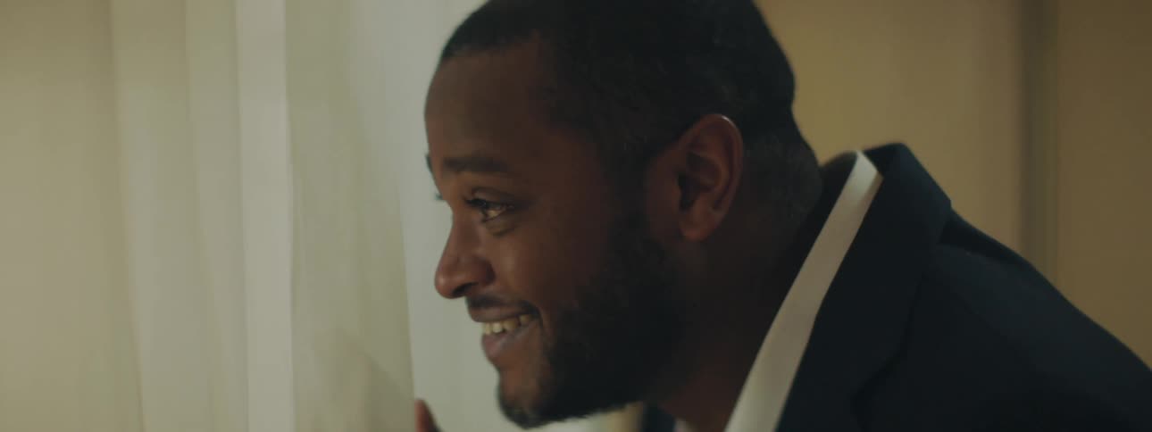 Man looks out of windows like he is watching someone and then smiles and waves and says something moving the curtains to be seen better