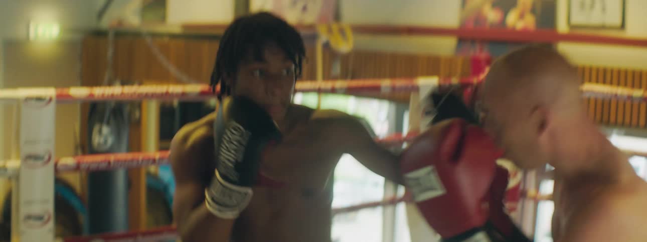 A pair of men exchange punches in a boxing ring in a small gym before one man knocks the other down with a hook