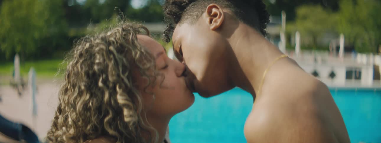 A teenage boy and girl kiss in front of a swimming pool