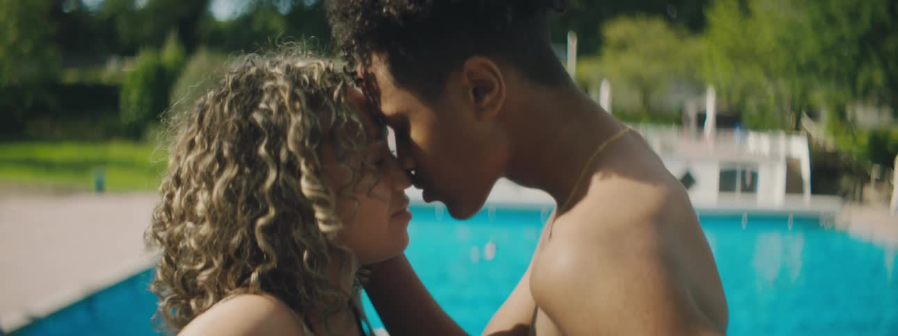 A teenage boy and girl kiss next to a swimming pool