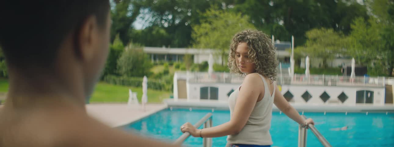 A teenager stands at the steps of a pool with a person swimming in it and looks back at a boy