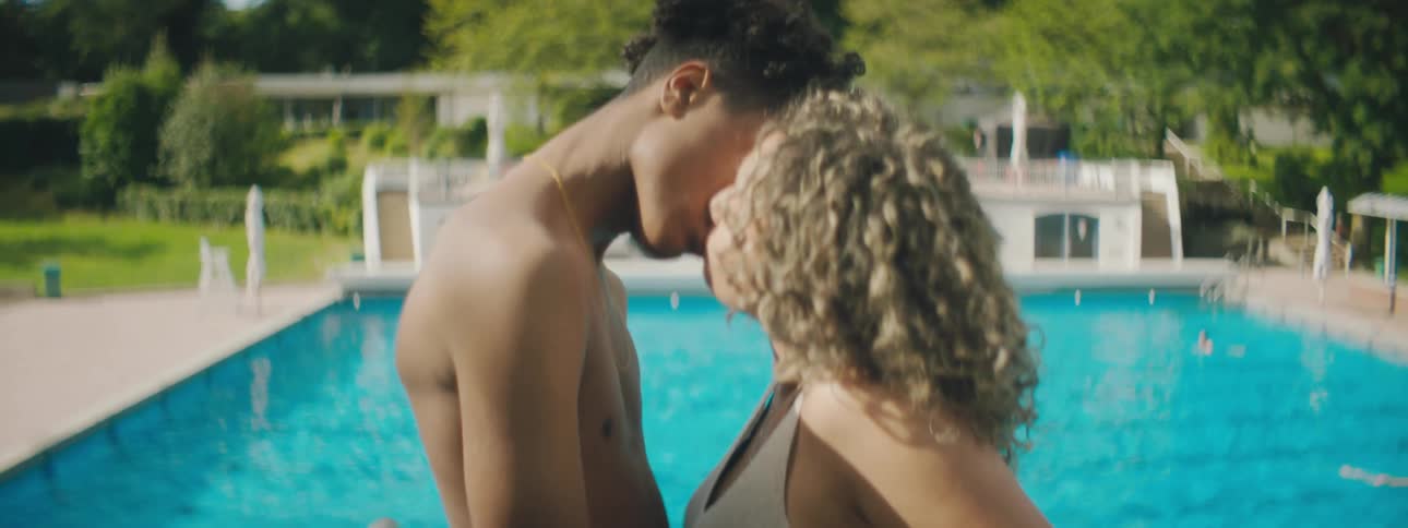 A teenage boy and girl kiss in front of a swimming pool
