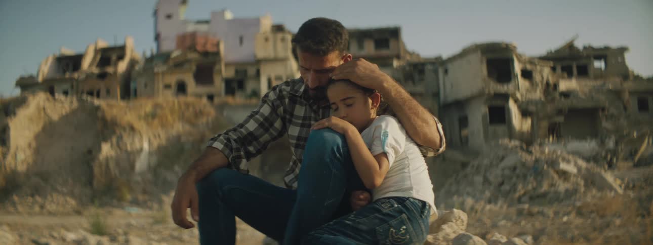 Father and son sit amongst piles of rubble with destroyed buildings and homes in the background