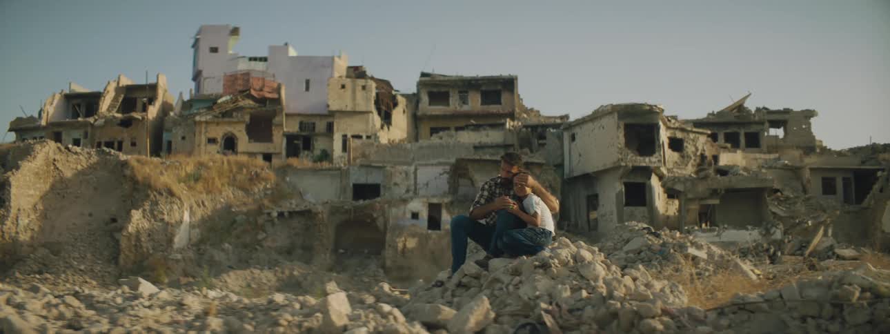 Father and son sit amongst rubble in front of destroyed buildings