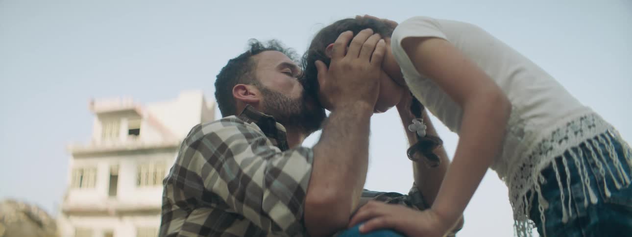 A girl approaches a depressed man sitting outdoors who kisses and cradles her head when she draws close
