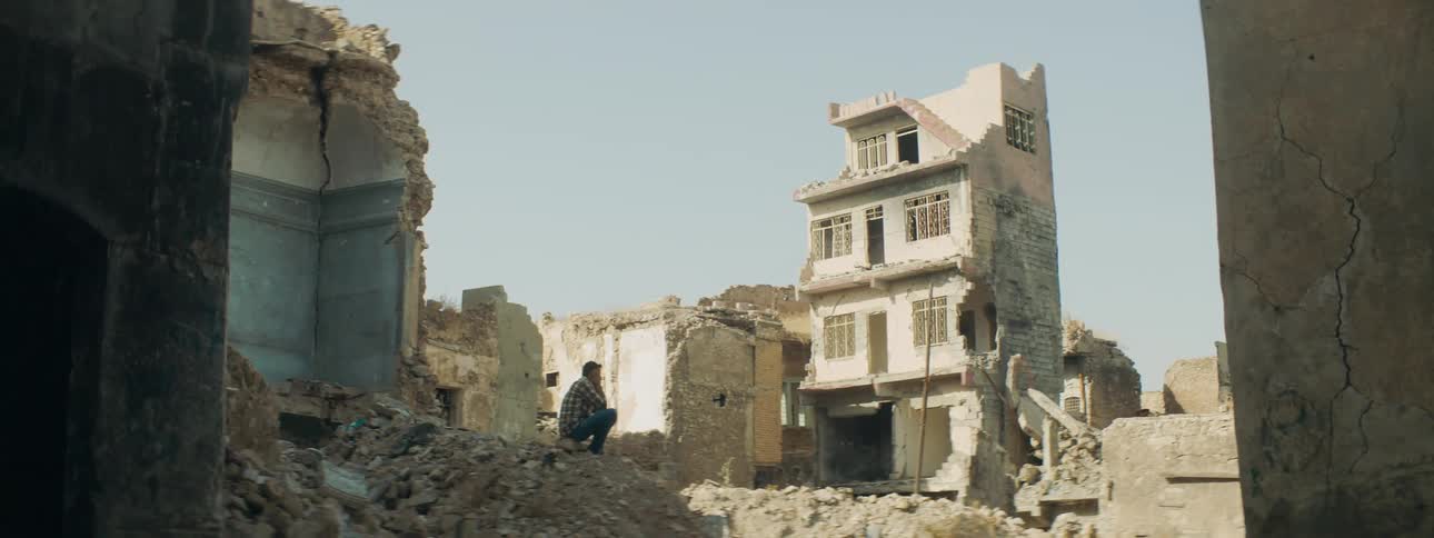 Man sits on rubble looking toward ruined building