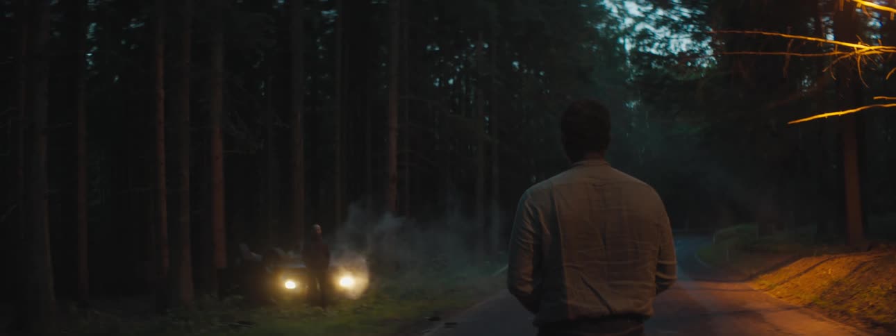A man stands on a road looking at another man standing in front his car that is off road in front of a forest