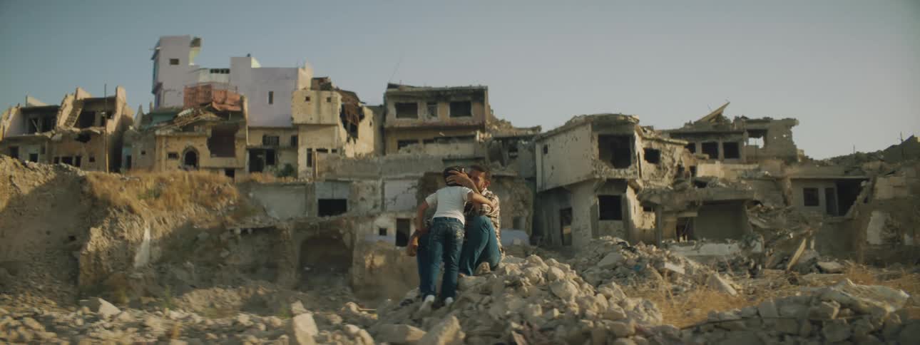 A father embraces his daughter while sitting on pile of rubble near abandoned town